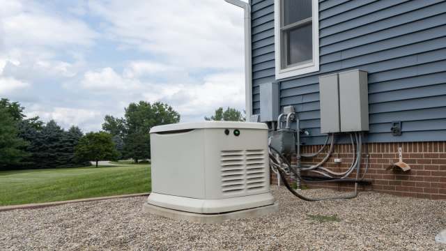 A generator is located beside a house, connected to electrical equipment and surrounded by gravel.