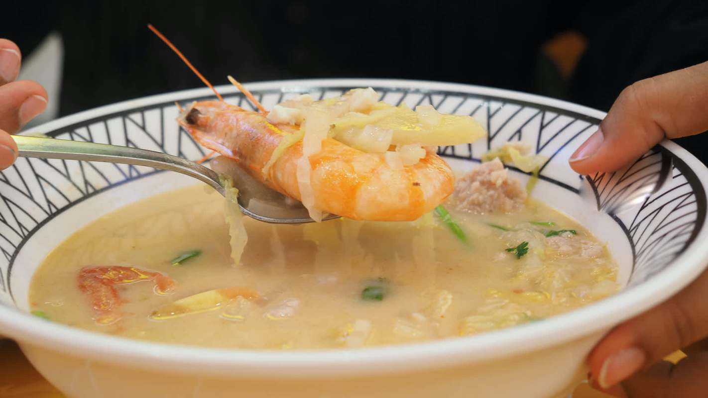 A person holds a spoon with shrimp and vegetables above a bowl of broth.