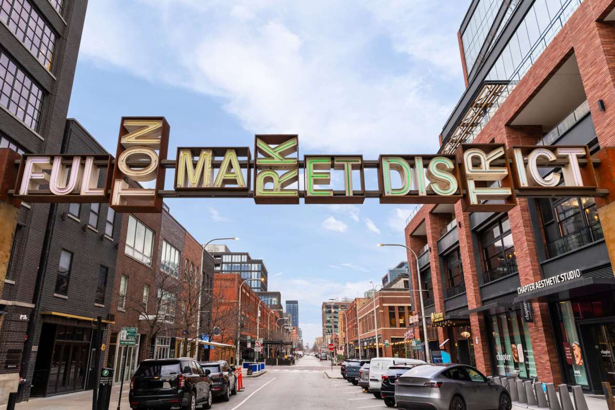 A street view of a market district with a sign overhead that reads "FULTON MARKET DISTRICT."