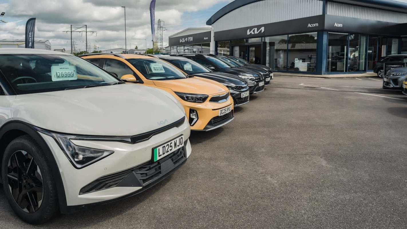 A row of various cars parked at a Kia dealership with price tags displayed.