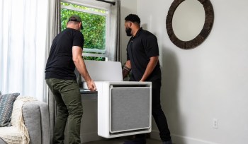 Two men are positioning a portable heat pump air conditioning unit near a window in a modern room.