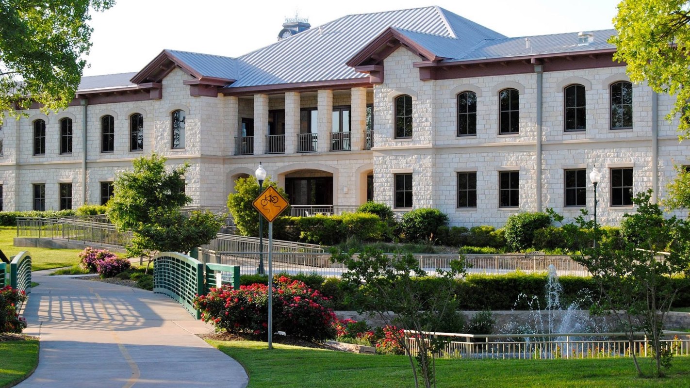A stone building surrounded by greenery and flowers, with a pathway and fountain in the foreground.
