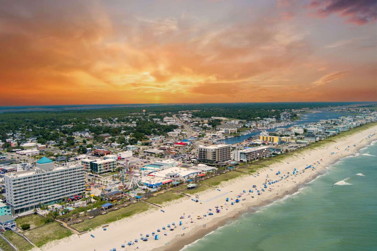 A coastal view of a beach with umbrellas, hotels, and a sunset in the background.