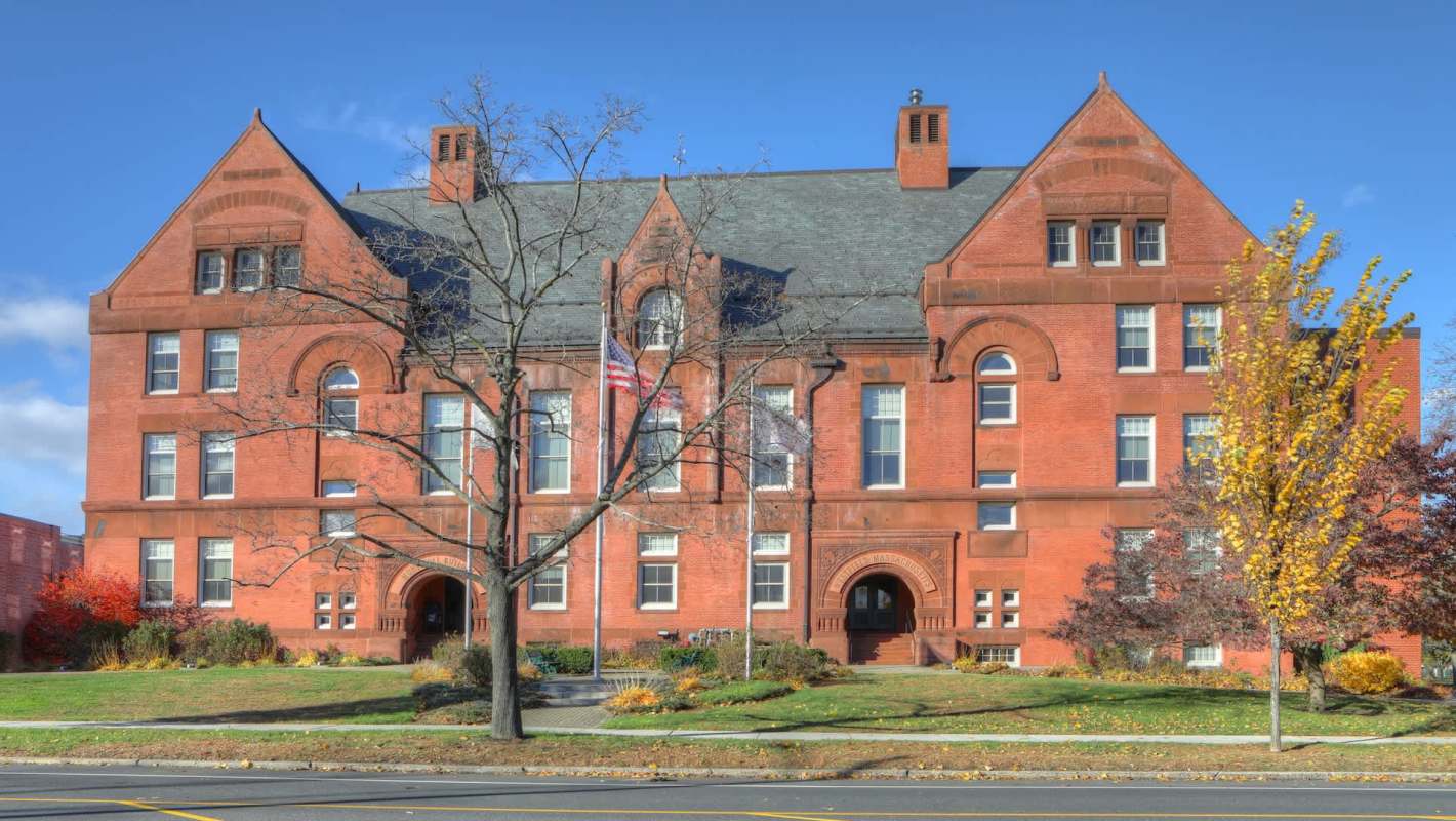 A red brick town hall with large windows and flags in front, surrounded by autumn trees and greenery.