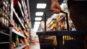 A hand holds a shopping basket in a grocery store aisle filled with packaged goods.