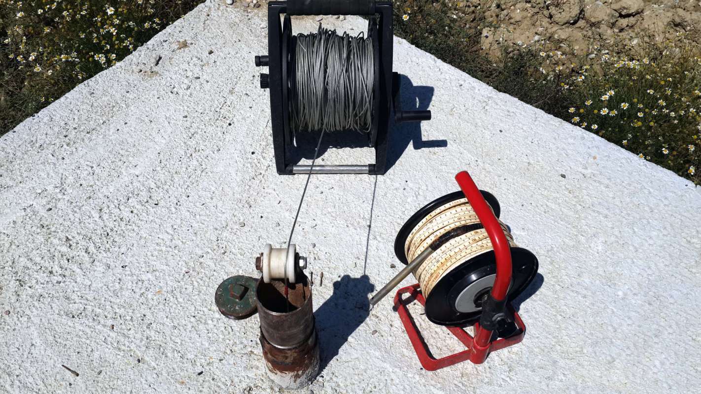 A metal spool with wire and a red reel with rope on a concrete surface next to a pipe and flowers.