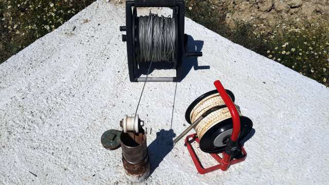 A metal spool with wire and a red reel with rope on a concrete surface next to a pipe and flowers.