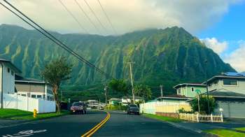 A road lined with houses and power lines, leading to steep green mountains under a cloudy sky.