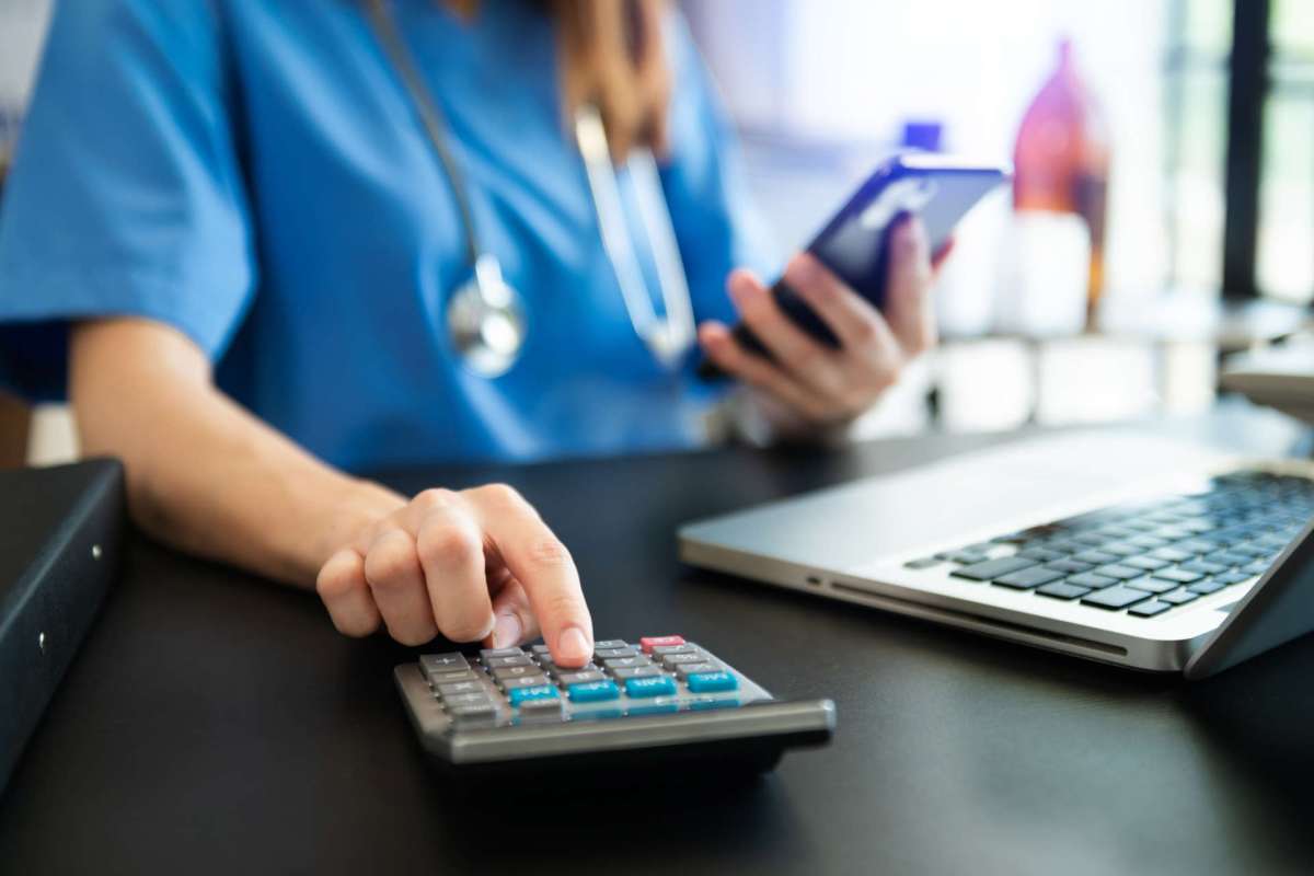 A person in scrubs uses a calculator while holding a smartphone next to a laptop on a desk.