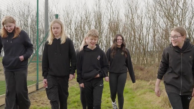 A group of five young people wearing black clothing walks along a grassy path with bare trees in the background.