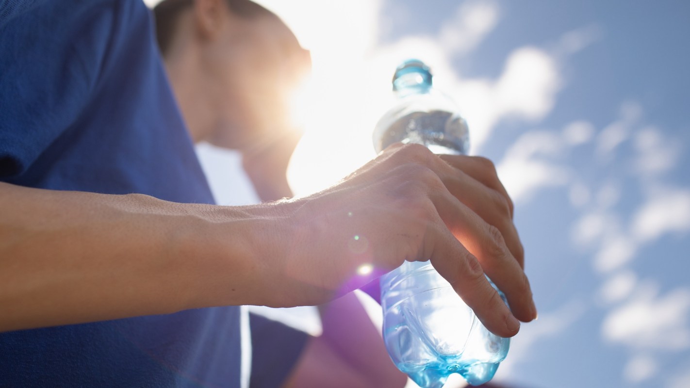 A person holds a water bottle while leaning against a railing under a bright sky.