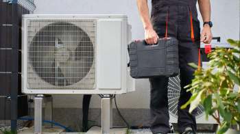 A technician holds a toolbox next to an outdoor air conditioning unit.