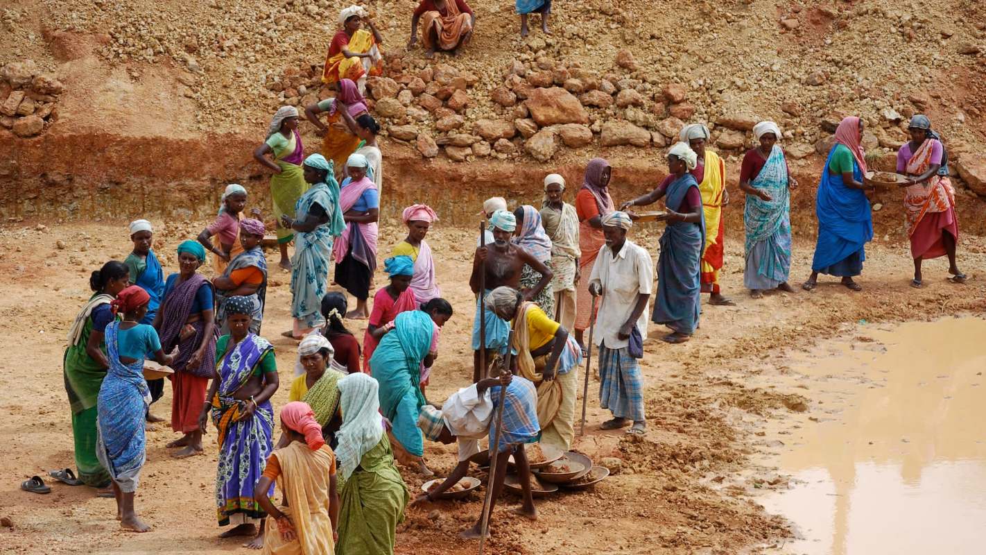 A group of people, mostly women in colorful saris, work together in a dirt quarry.