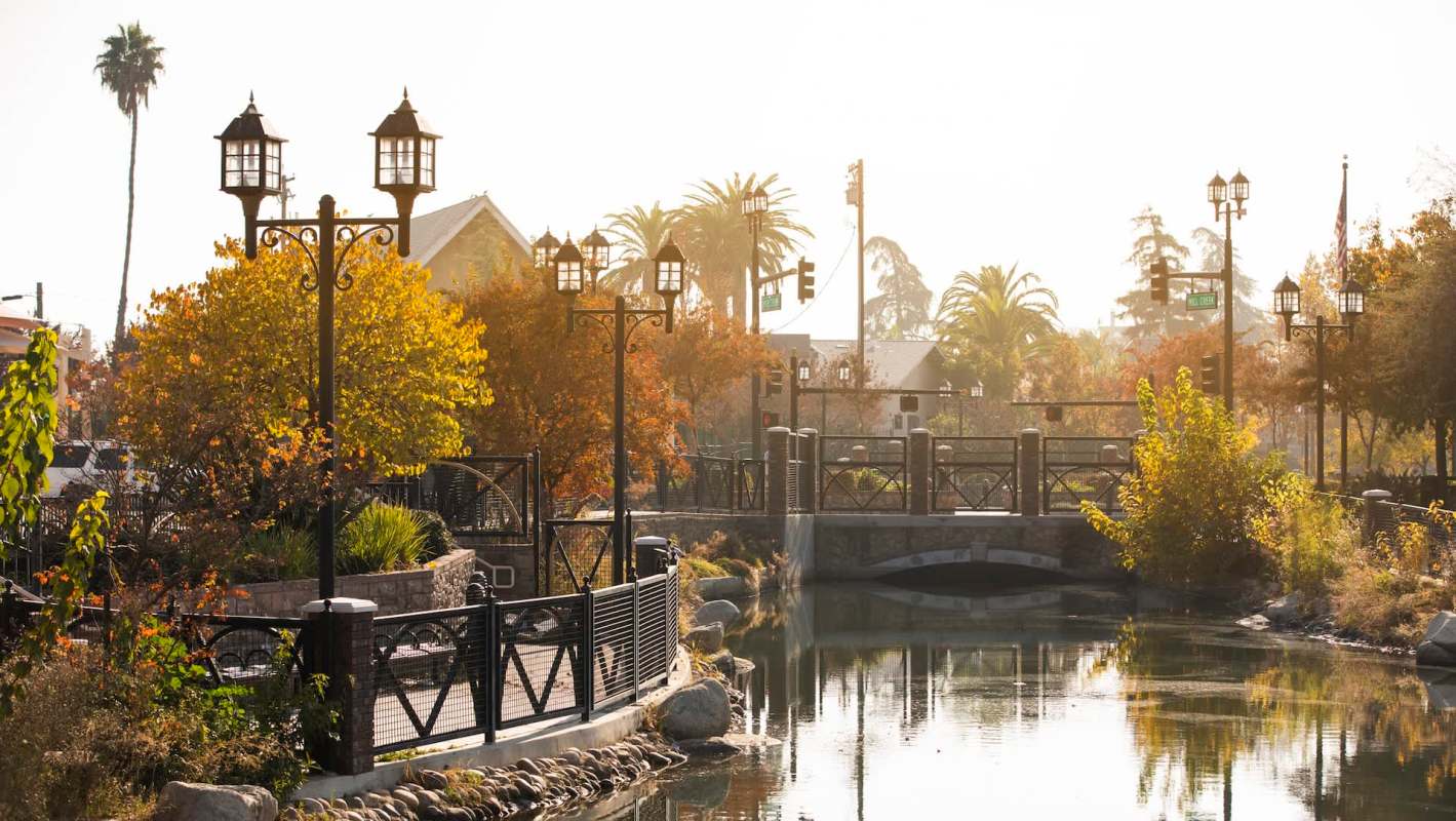 A park scene under bright sunlight featuring a pond, bridge, trees with autumn leaves, and decorative lamp posts.