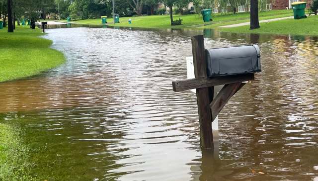 A mailbox stands in a flooded residential area with standing water and green lawns.
