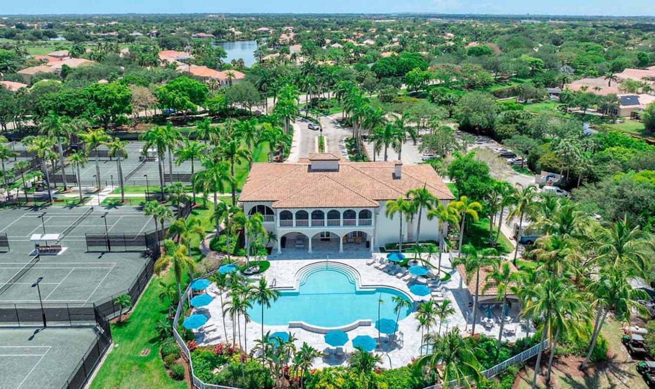 Aerial view of a homeowners association complex with a pool, palm trees, and adjacent tennis courts in a landscaped area.
