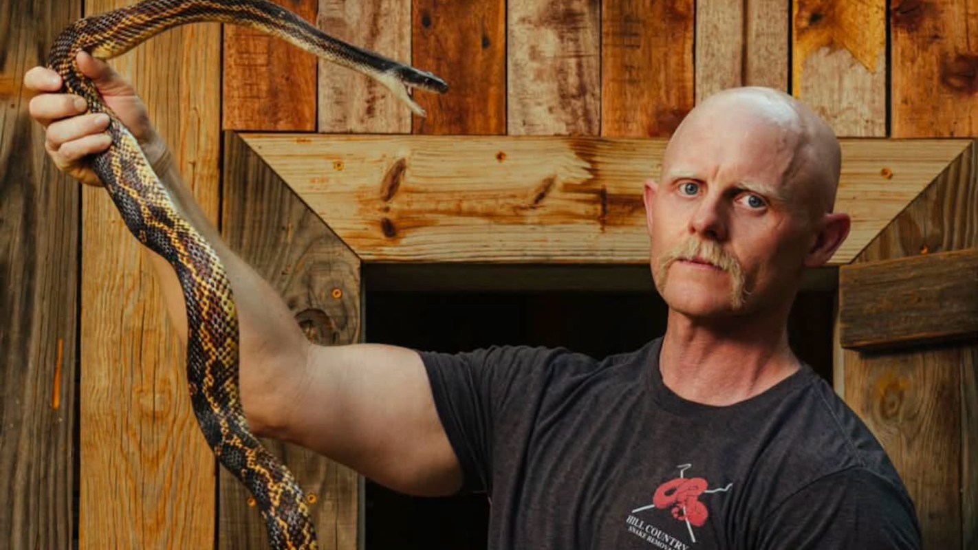 A person holds a large snake in front of a wooden background.