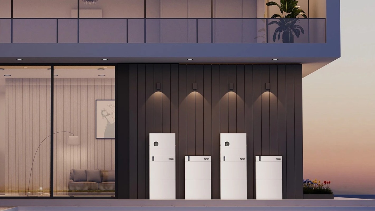 Four modern white refrigerators are lined up against a dark wooden wall, with a glass patio in the background.