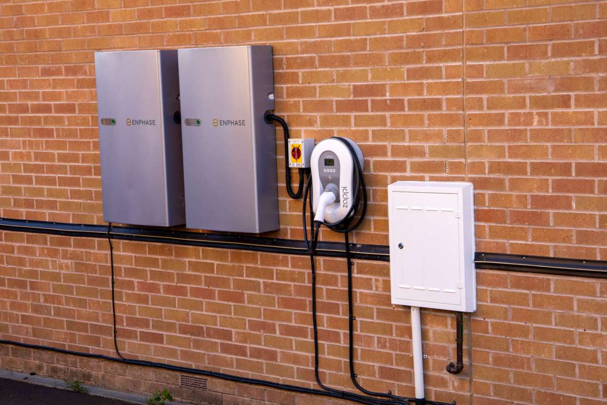 Two gray energy storage units mounted on a brick wall with an electric vehicle charging station and an electrical box.