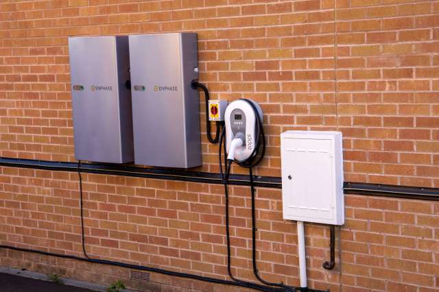 Two gray energy storage units mounted on a brick wall with an electric vehicle charging station and an electrical box.