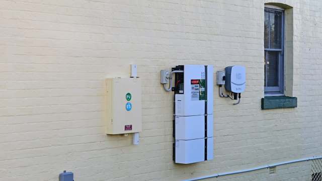 Electrical equipment and inverters mounted on a yellow brick wall beside a window.