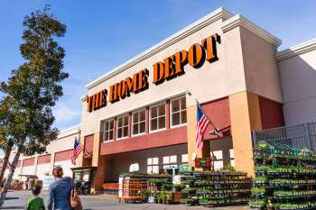 A Home Depot store exterior with plants for sale and an American flag displayed.