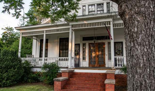 A large white house with a porch, steps, and greenery surrounding it.