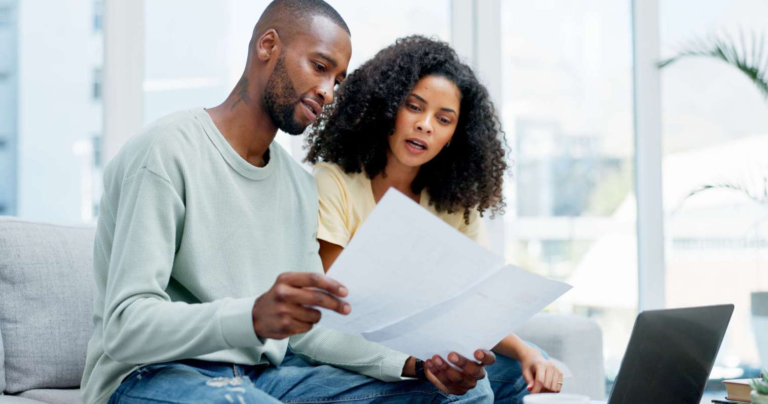 A man and a woman sit on a sofa, reviewing documents with a laptop nearby.