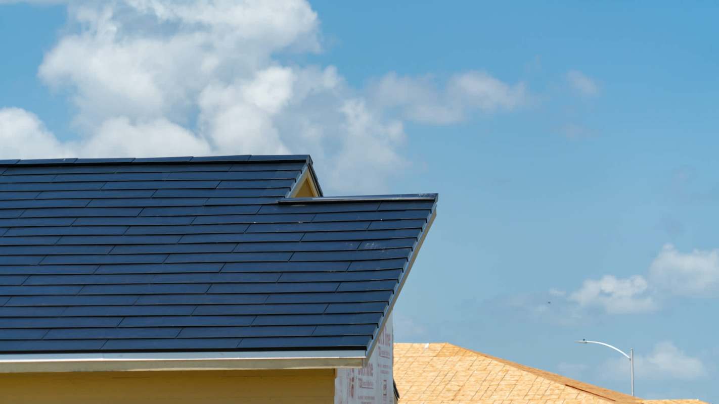 A rooftop with dark shingles against a blue sky with fluffy clouds.