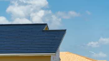 A rooftop with dark shingles against a blue sky with fluffy clouds.