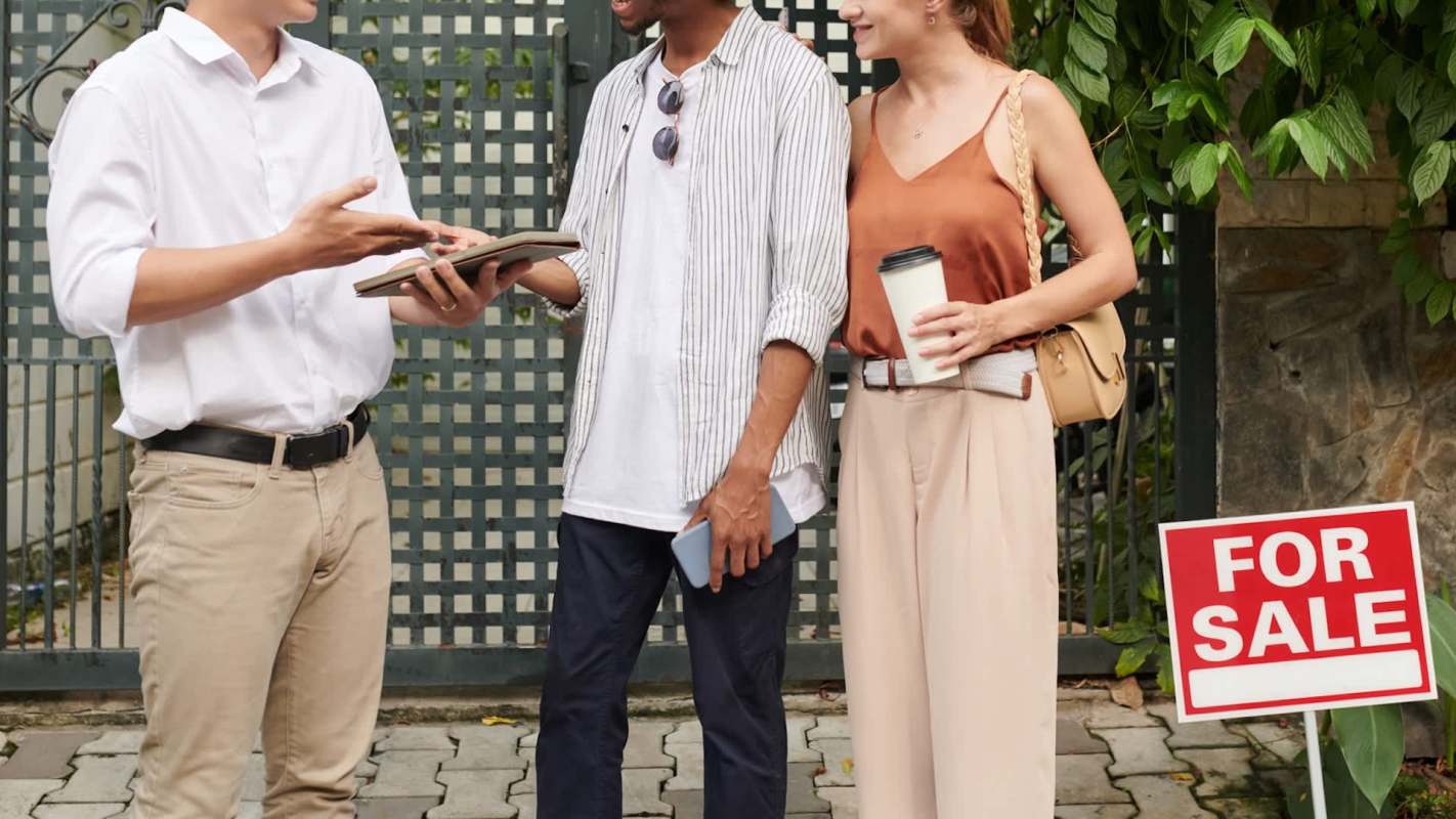 Three people converse in front of a house with a "For Sale" sign.
