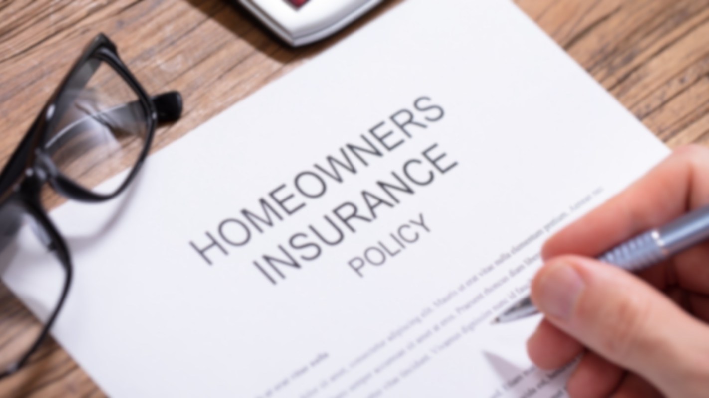 A hand holding a pen above a homeowners insurance policy document with glasses nearby.