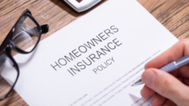 A hand holding a pen above a homeowners insurance policy document with glasses nearby.