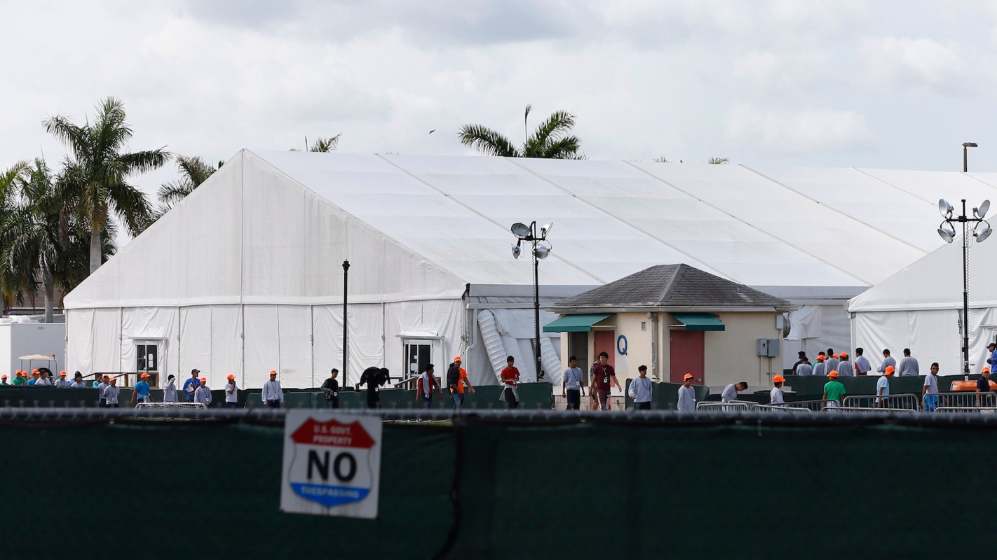 A large white tent with people wearing orange hats near a fenced area and palm trees in the background.
