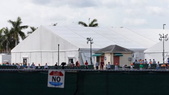 A large white tent with people wearing orange hats near a fenced area and palm trees in the background.