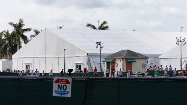 A large white tent with people wearing orange hats near a fenced area and palm trees in the background.