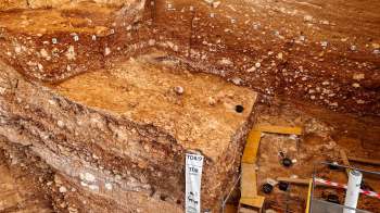 An archaeological excavation site with layers of earthy sediment, measuring tools, and excavation equipment.