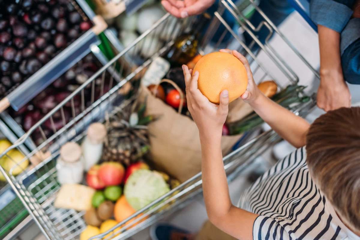 A child holds a grapefruit while standing near a shopping cart filled with various grocery items.