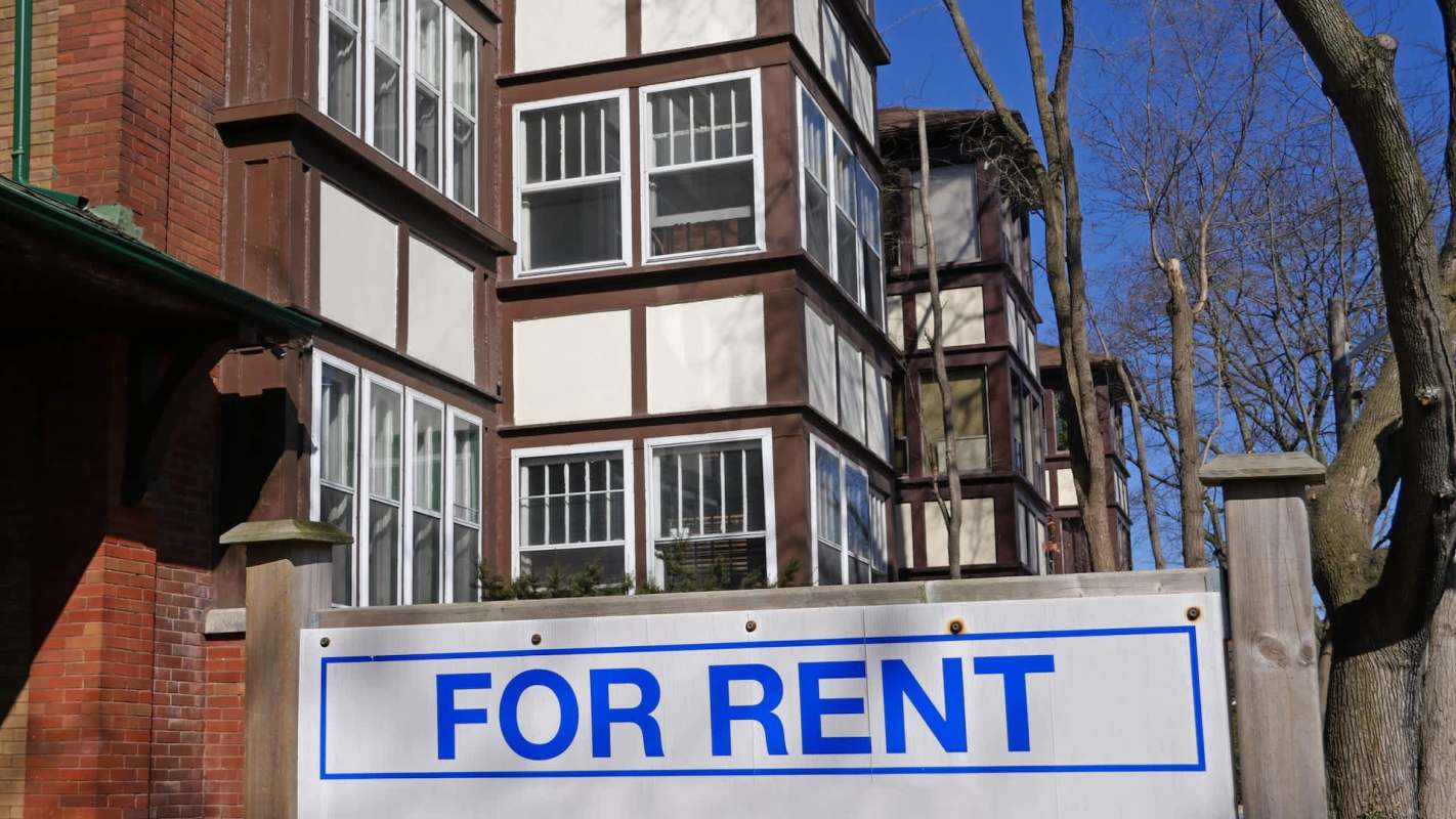 A rental sign in front of a multi-story building with a mix of brick and wood siding.