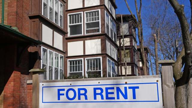 A rental sign in front of a multi-story building with a mix of brick and wood siding.