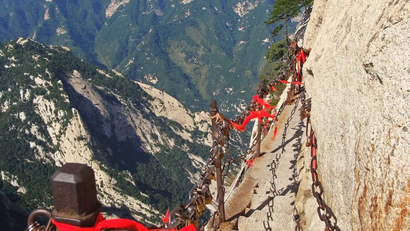 A narrow path along a mountain edge with chains and red ribbons, overlooking steep, green hills.