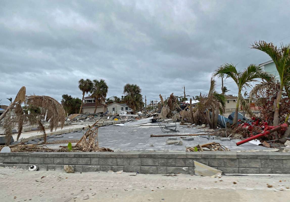 A damaged area with debris, downed palm trees, and destroyed buildings under a cloudy sky.