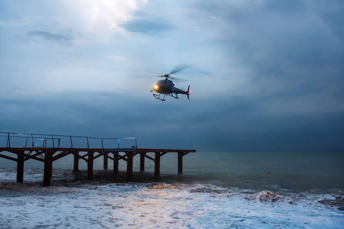 A helicopter hovers above a pier over turbulent ocean waters under a cloudy sky.