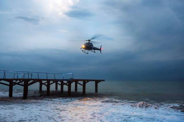 A helicopter hovers above a pier over turbulent ocean waters under a cloudy sky.