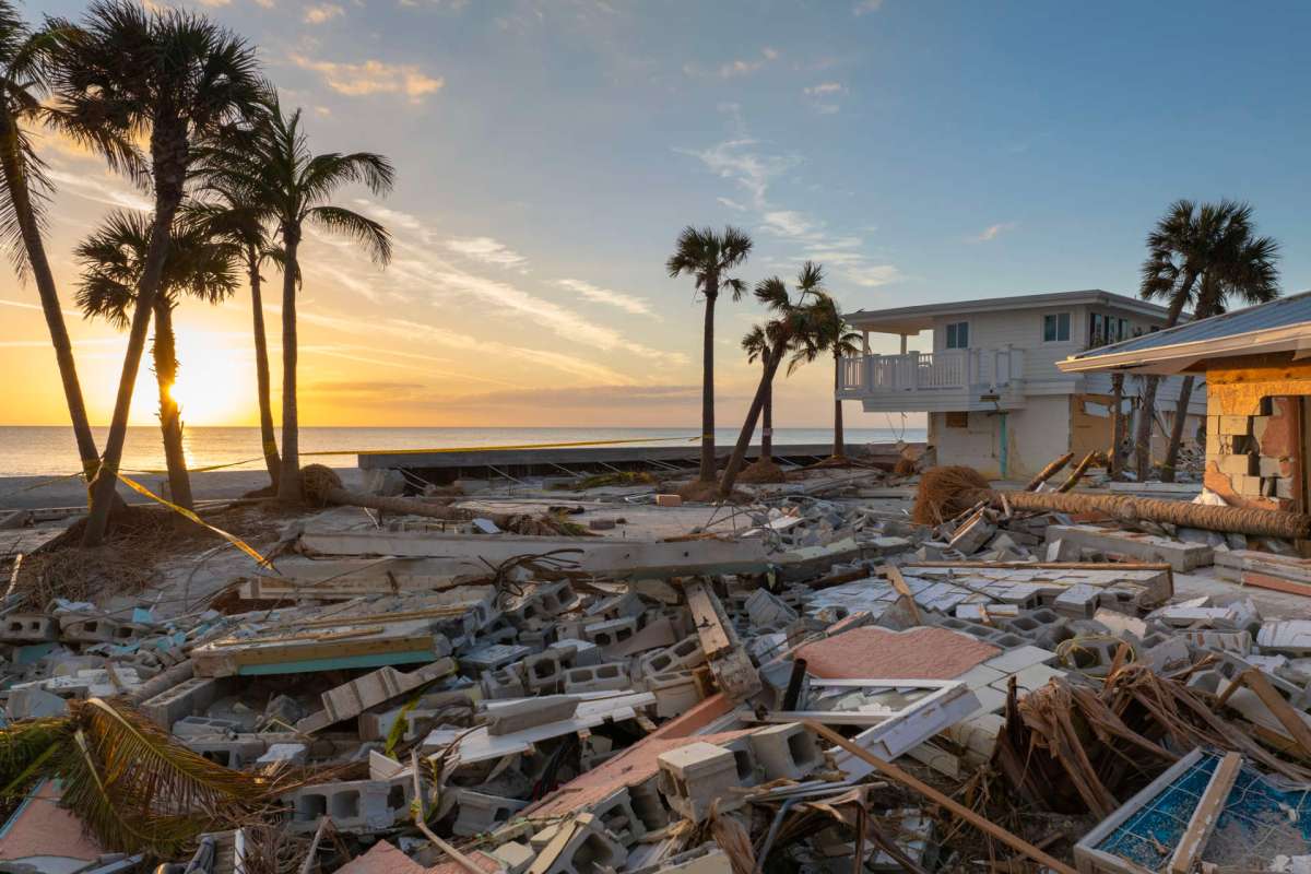 A landscape of destruction with debris, palm trees, and a house near the beach at sunset.