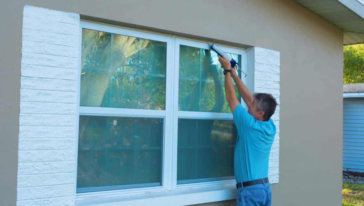 A person applies caulk around a window frame on a house.