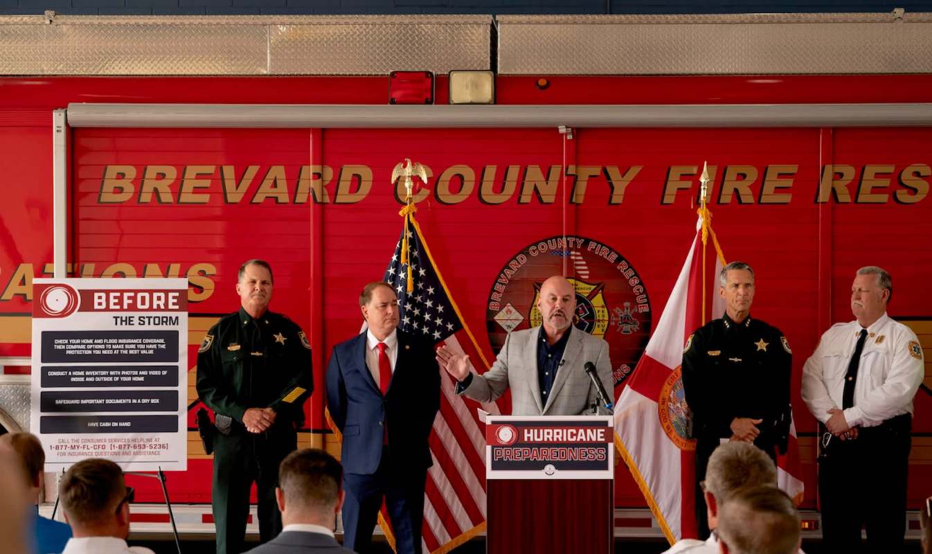 A man speaks at a podium during a press conference on hurricane preparedness, with officials standing nearby.