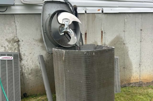 An air conditioning unit with an exposed fan and condenser coils next to a concrete wall.