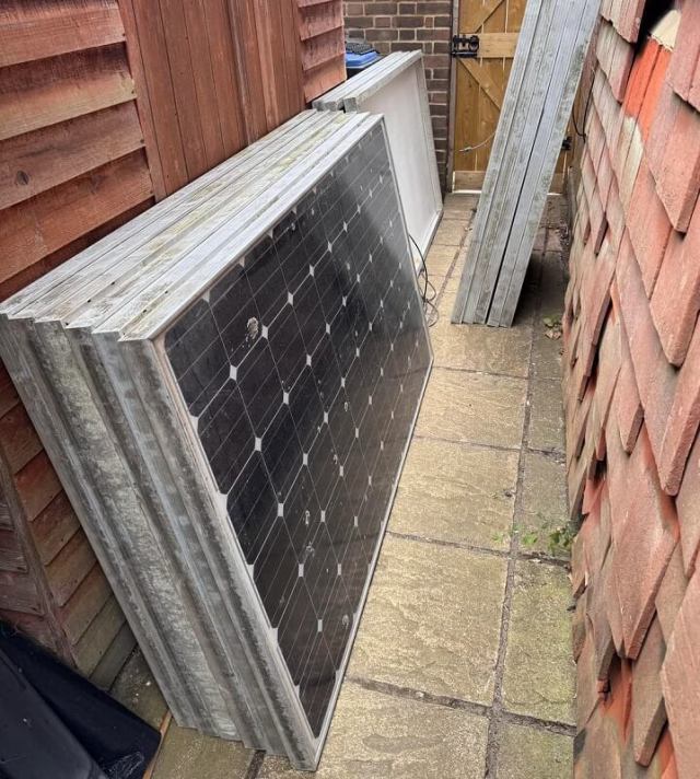 Solar panels leaning against a wooden fence in a narrow outdoor space.