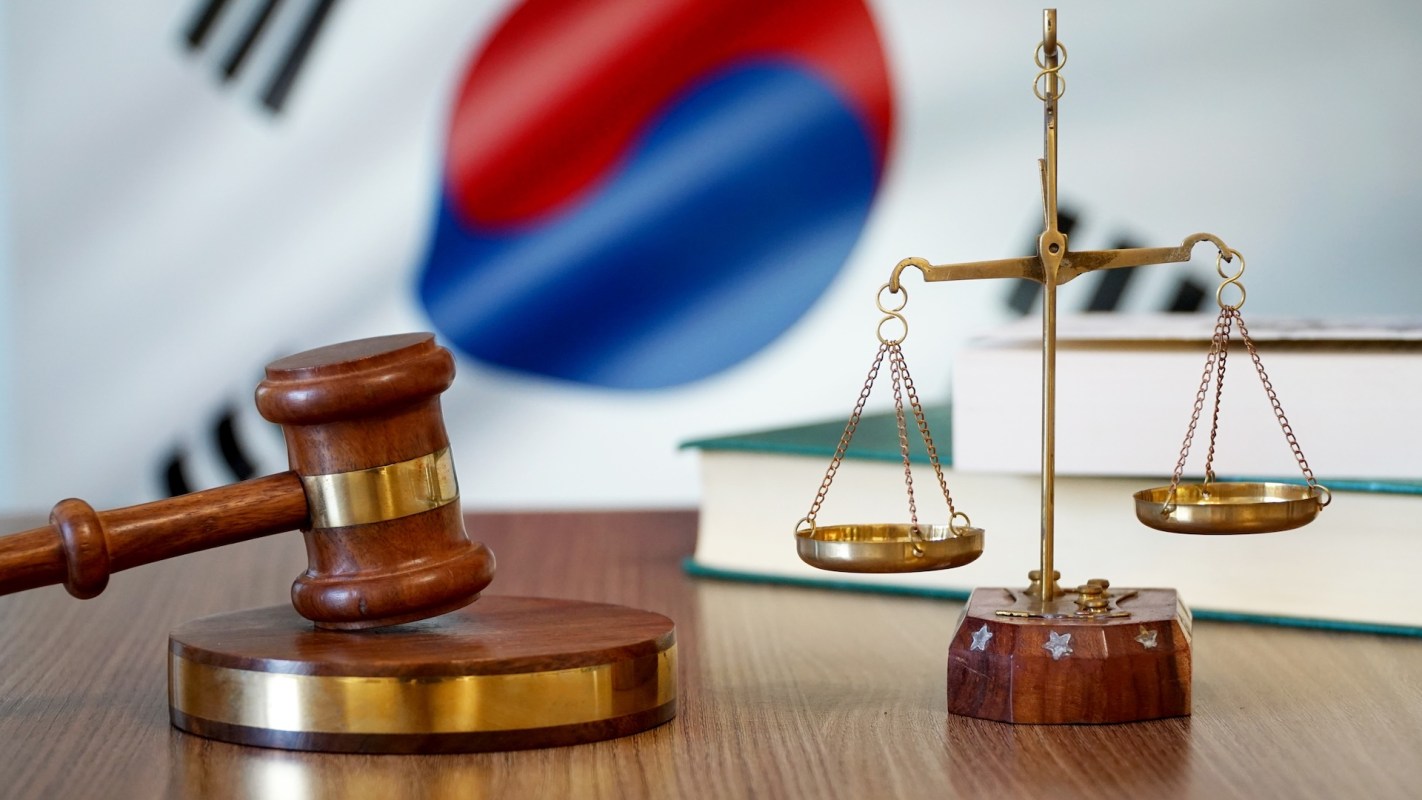 A gavel and balance scales on a table with a blurred South Korean flag in the background.
